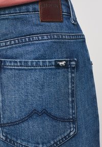 Vaqueros de mezclilla azul claro con una etiqueta de cuero marrón con la inscripción "MUSTANG." Presentan un patrón de costura ondulada distintivo y una pequeña etiqueta de logo en el bolsillo trasero.