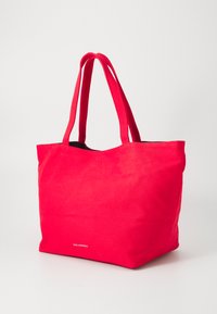 Sac fourre-tout en toile rouge avec deux longues poignées, ouverture supérieure et intérieur noir. Présente un logo discret en bas sur le devant.