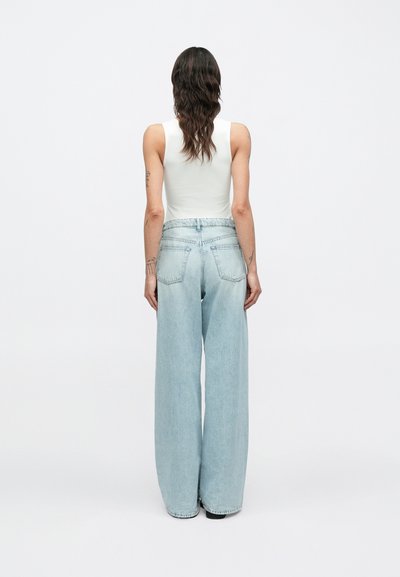 Φαρδιά τζιν σε ανοιχτό μπλε χρώμα με ψηλή μέση και δύο πίσω τσέπες, σε συνδυασμό με λευκό αμάνικο τοπ, από πίσω.