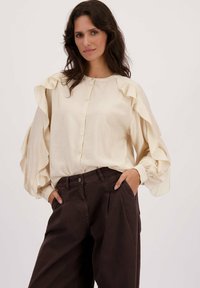 Femme portant un chemisier crème à boutons avec des manches volantes et un pantalon large taille haute sombre, les mains dans les poches, fond neutre.