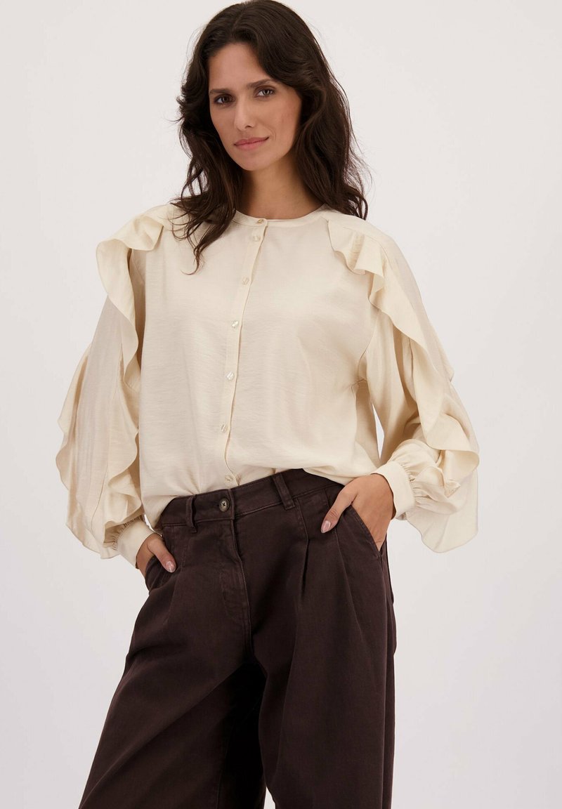 Femme portant un chemisier crème à boutons avec des manches volantes et un pantalon large taille haute sombre, les mains dans les poches, fond neutre.