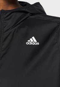 Musta tekstuuriga Adidas kapuutsiga jakk, millel on valge Adidas logo rinnal, kandmas isik, kes näitab ülemist kehaosa ja kaela.