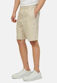 Pantaloni corti in cotone beige con cintura elastica, tasche laterali e taglio dritto, abbinati a sneakers bianche.