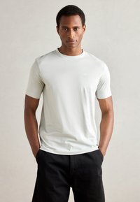 Lichtgrijs t-shirt met korte mouwen, gemaakt van gladde stof, met een ronde halslijn en een subtiel logo op de borst. Gecombineerd met zwarte broek.