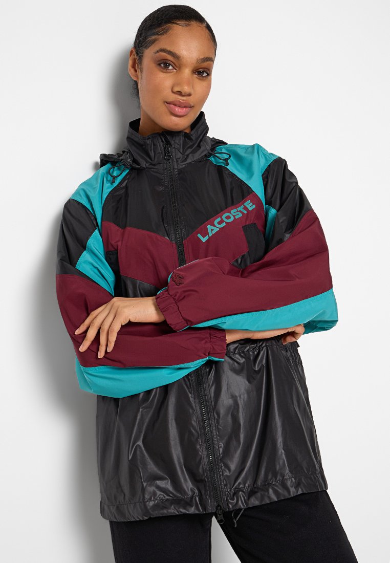 Kurtka przeciwwiatrowa Lacoste w kolorach czarnym, burgundowym i teal. Posiada zamek z przodu, elastyczne mankiety oraz design w blokach kolorowych z akcentem w postaci logo.