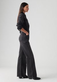 Juodas džinsinis kombinezonas su ilgomis rankovėmis, šoninėmis kišenėmis, plačiomis kelnėmis ir prigludusia liemene. Lygus paviršius su šiek tiek nublukusiu apdaila.