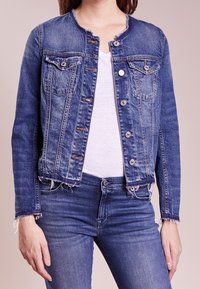 Jeansjacka i medelblått med rundad hals, knappstängning framtill, två bröstfickor och slitna kantdetaljer. Bärs över en vit skjorta.