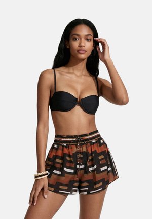 Donna che indossa un top bikini nero e pantaloncini marroni, neri e bianchi con fantasia e coulisse con braccialetti a un polso su sfondo semplice.