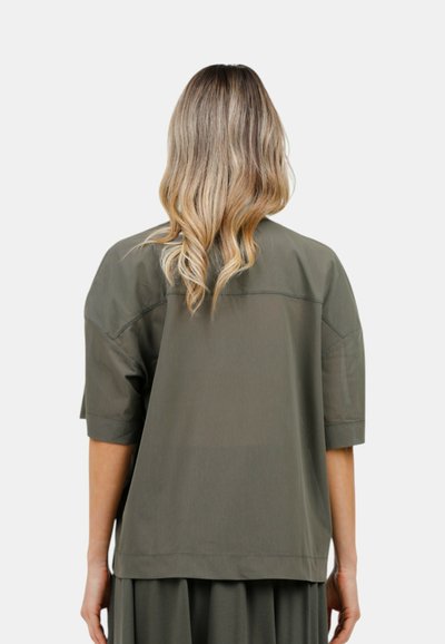 Camicia over size verde oliva, maniche corte, tessuto morbido, vestibilità rilassata, cuciture decorative sulle spalle, abbinata a una gonna coordinata.