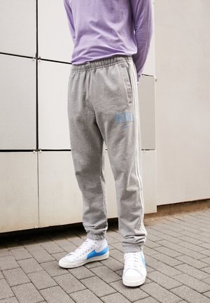 Personne portant un pantalon de jogging Nike gris, des baskets blanches avec un swoosh bleu, et un t-shirt à manches longues violet clair, debout sur le trottoir.