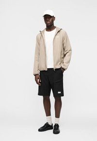 Moški model v bež jakni z zadrgo, beli majici, črnih kratkih hlačah, belih nogavicah, črnih supergah in beli kapici, stoji z eno roko v žepu.