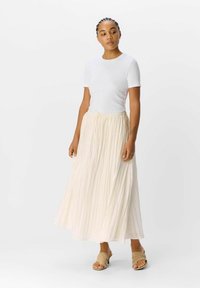 Top corto de manga corta blanco con textura de canalé, combinado con una falda larga de color beige claro y pliegues, que presenta una cinturilla elástica y cordón ajustable. Sandalias en tonos neutros.