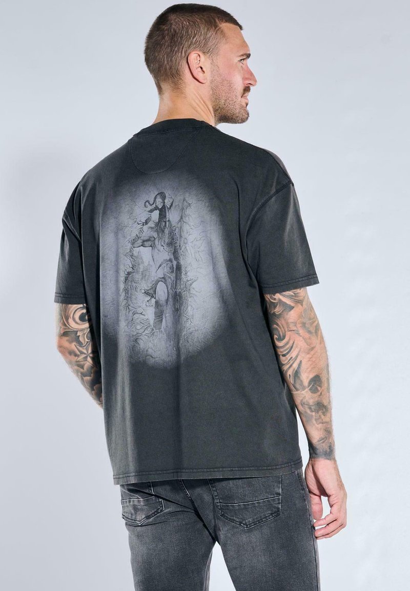 Man with tattooed arms wearing black t-shirt with faded graphic of warrior and child on back, paired with dark gray jeans.