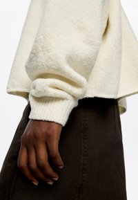Cremfarbener Fleece-Pullover mit gerippten Bündchen, getragen über einem dunkelbraunen Kleidungsstück. Die Textur ist weich und leicht flauschig.