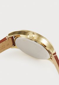 Montre en acier inoxydable doré avec un boîtier lisse et un bracelet en cuir marron. Elle dispose d'un cadran rond et d'une couronne latérale en accent.