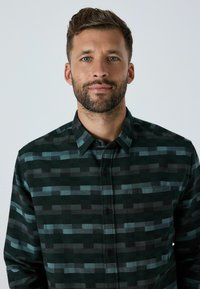 Napapaita tummanvihreästä ja mustasta ruutukuviosta. Varustettu rintataskulla ja kauluskannalla. Valmistettu teksturoidusta kankaasta.