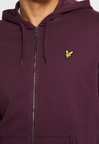 Person som bär en lila hoodie med dragkedja och en gul fågel-emblem på vänster bröst, samt snoddar som hänger från huvan.