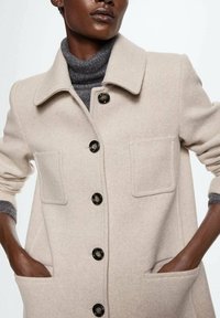Beige uldfrakke med krave, frontlomme og sorte knapper. Det indvendige lag er en grå rullekrave sweater.