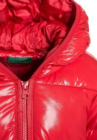Röd puffjacka med glänsande yta, har dragkedja, elastiska ärmslut och fodrad huva. Etikett synlig inuti med varumärkesnamn.