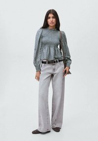 Blusa verde a fantasia con corpo arricciato e maniche lunghe, abbinata a jeans grigi chiari a zampa d'elefante e scarpe slip-on scure a fantasia.