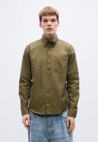 Πουκάμισο σε ελιά πράσινο, βαμβακερό ύφασμα, τσέπη στο στήθος, ταμπελάκι με λογότυπο, μακριά μανίκια, συνδυασμένο με ανοιχτό μπλε τζιν με φθορές.