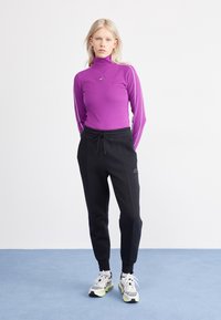 Långärmad lila topp med hög krage och vita randiga detaljer, som matchas med svarta sweatpants med dragsko och Nike-logga. Vita och grå sneakers.