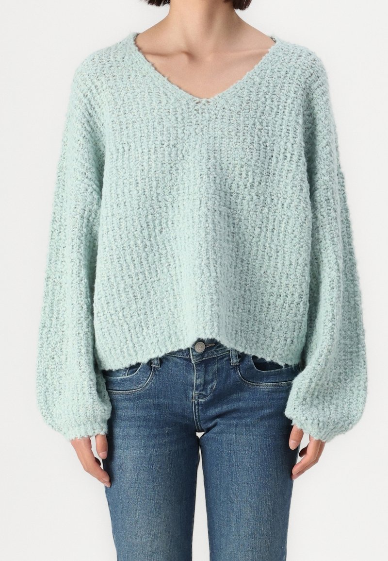 Personne portant un pull en maille texturée bleu clair avec de longues manches bouffantes et un jean bleu ajusté sur un fond uni.