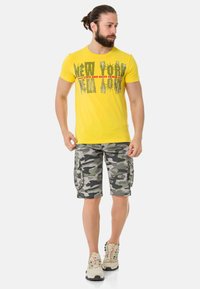 Gelbes Baumwoll-T-Shirt mit schwarzem Grafiktext und roten Akzenten, kombiniert mit graugrünen Camouflage-Cargo-Shorts. Eine lässige Schuhmode vervollständigt den Look.