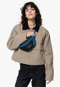 Mustat kangastaskulaukku sinisillä yksityiskohdilla ja vetoketjulla, joka on pidetty vartalon ympäri. Beige ylisuuressa takissa, jossa on corduroy-kaulus, ja tummissa farkuissa.
