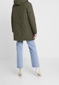 Olivově zelený prošívaný kabát s kapucí, rovného střihu a střední délky, doplněný světle modrými džíny se širokými nohavicemi a béžovými kozačkami na podpatku.