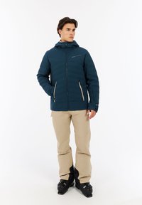 Námornícky izolačný kabát s rozopínateľnými vreckami, predným zipsom a textúrovaným povrchom. Nosený s béžovými nohavicami a čiernymi lyžiarskymi topánkami v stojacej póze.