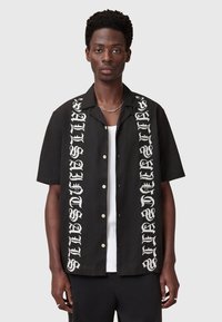 Chemise noire à manches courtes avec du texte graphique blanc sur les deux côtés, fermeture à boutons à l'avant et coupe décontractée. Fabriquée en tissu léger.