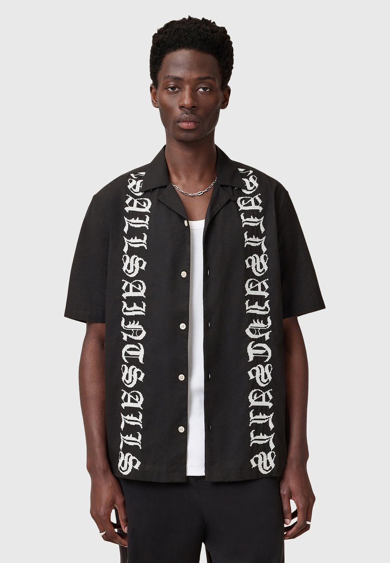 Chemise noire à manches courtes avec du texte graphique blanc sur les deux côtés, fermeture à boutons à l'avant et coupe décontractée. Fabriquée en tissu léger.
