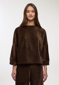 Top marrone a maniche lunghe in tessuto vellutato con vestibilità ampia, polsini larghi e collo leggermente alto, abbinato a pantaloni marroni coordinati.