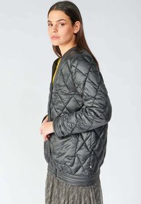 Veste en nylon noir matelassée avec un col rond, des manches longues, deux poches avant et des poignets côtelés, mise en valeur par une doublure intérieure jaune.