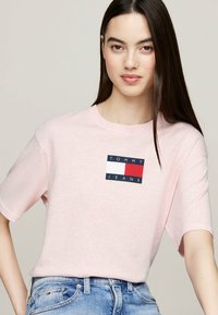 Camiseta de manga corta rosa hecha de tela suave, con un parche de logo 'TOMMY JEANS' en azul, blanco y rojo en el pecho. Diseño casual.