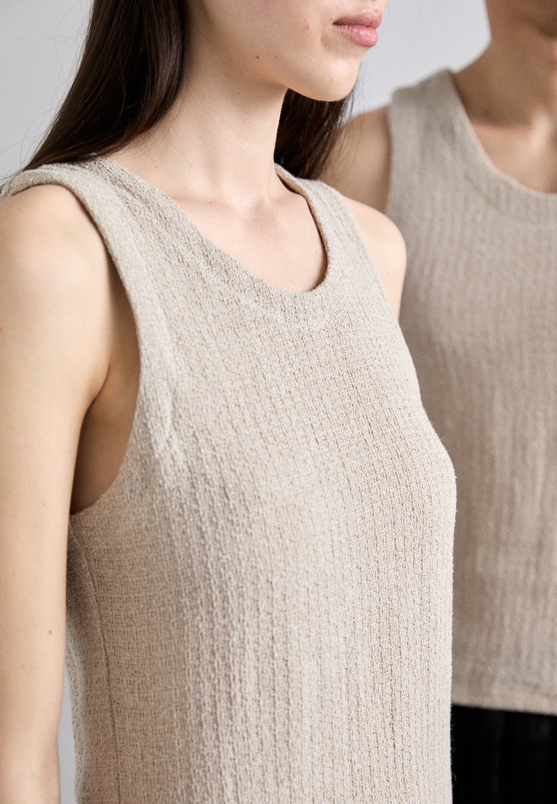 Beige ärmelloses Top mit gerippter Textur, rundem Ausschnitt und breiten Trägern; der Stoff wirkt weich und leicht dehnbar.