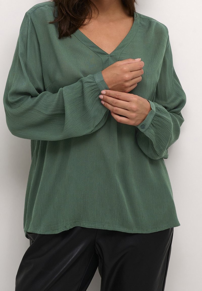 Femme ajustant le poignet d'un chemisier vert ample à manches longues porté avec un pantalon noir sur un fond uni.