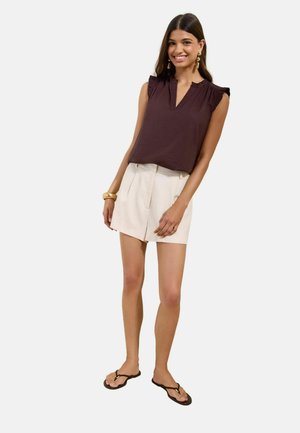 Jonge vrouw die lacht, draagt een mouwloze donkerbruine blouse, lichtbeige shorts, sandalen en oorbellen en armband in goudkleur, staand op een witte achtergrond.