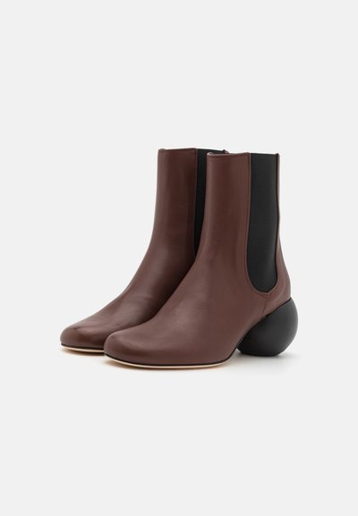 Bottines en cuir marron avec un talon noir arrondi, des panneaux latéraux élastiques et un design épuré, présentant une texture lisse et un bout pointu.