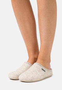 Chaussons en feutre gris clair avec un design à bout fermé, bord cousu et petite étiquette de marque en métal. Texture douce et style facile à enfiler.