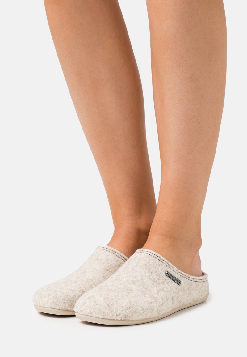 Chaussons en feutre gris clair avec un design à bout fermé, bord cousu et petite étiquette de marque en métal. Texture douce et style facile à enfiler.
