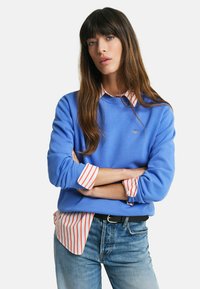 Felpa blu con polsini a coste, indossata sopra una camicia a righe rosse e bianche con colletto. Il modello indossa jeans azzurri e una cintura nera.