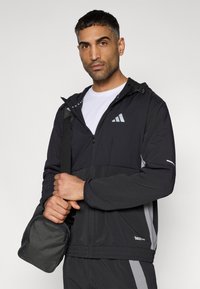 Μαύρο μπουφάν με φερμουάρ και ανακλαστικές λεπτομέρειες, ριμπ μανσέτες και ρυθμιζόμενο κουκού λι, φορεμένο πάνω από λευκό μπλουζάκι. Κρατείται μια μικρή μαύρη τσάντα.