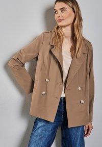 Giacca beige doppiopetto con grandi bottoni argentati, vestibilità su misura, rever a punta e maniche lunghe, indossata sopra un top a scollo a V chiaro.