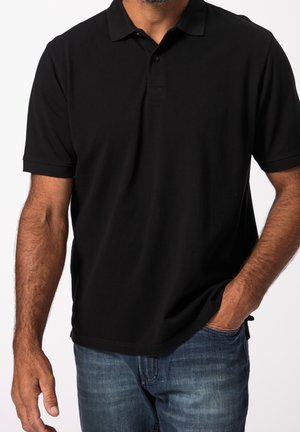 Man wearing a black short-sleeve polo shirt with one hand in the pocket of blue jeans against a white background.