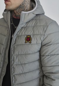 Szara kurtka puchowa z kapturem, z pikowaną fakturą i zapięciem na zamek. Wyszywane logo po lewej stronie klatki piersiowej; widoczna warstwa z dzianiny ściągaczowej pod spodem.