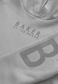 Gaiši pelēks džemperis ar V veida kakla izgriezumu. Iezīmēts ar teksturētu drukājumu "BAKER by TED BAKER" un lielu burtu "B" uz piedurknes. Mīksta auduma.