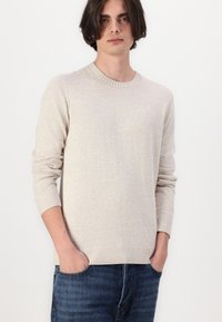 Világos bézs kötött pulóver, kör alakú nyakkivágással, bordázott mandzsettákkal és aljjal. Kék farmernadrág fölött viselve, amely laza szabású.