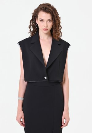 Donna con capelli ricci che indossa un blazer nero corto senza maniche con rever a incavo e bottone decorativo, gonna nera, orecchini a cerchio e braccialetto d'argento.